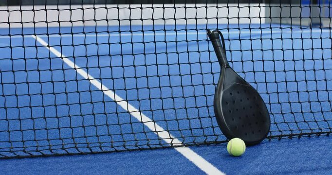 Padel racket and ball resting against net on blue court, copy space
