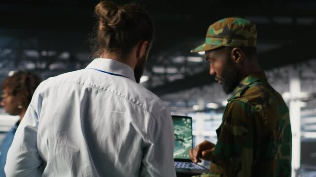 Military technician reviewing tactical dashboard on laptop screen inside secure server hub. Army expert using strategic communications and infrastructure oversight software on notebook, camera B