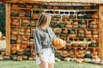 young woman stands at pumpkin festival, holding large orange and green pumpkin. She is surrounded by rows of pumpkins and a wooden display. The autumn farm setting captures the harvest season vibe.