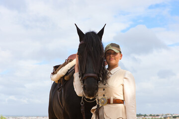 Obraz premium Young and beautiful woman holds with affection the face of her black purebred Spanish horse and looks at the camera with happiness. The woman is dressed in typical horsewoman costume.