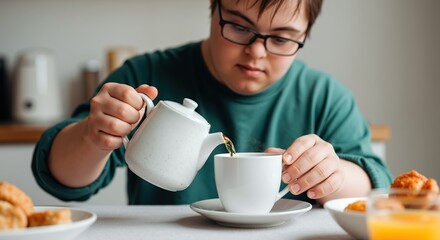 Young man with down syndrome wearing glasses pouring tea from teapot into cup at home. Person with special needs enjoying hot beverage in kitchen. Inclusion, independent living. Home care service sale