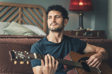 Young man sitting on bedroom floor playing acoustic guitar, gazing thoughtfully into distance, representing creativity, emotion and lifestyle music moments at home., man relaxed holiday at home