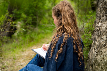 Unrecognizable woman with curly hair writing something in notebook outdoors relaxing spending...