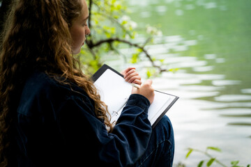 Rear view of calm woman with curly hair writing thoughts in notebook by lake relaxing spending leisure time alone keeping diary journaling dreassed casual dark blue shirt sitting in nature outdoors