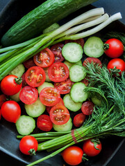 Fresh red cherry tomatoes, green leek, radish and cucumber in pan on the black background, top view of vegetables covered with water drops.