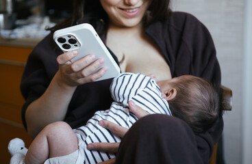 Beautiful young woman is breastfeeding her baby boy at home, close-up view. She is working online at the same time, remote work from home. Chatting with friend, having online shopping. 
