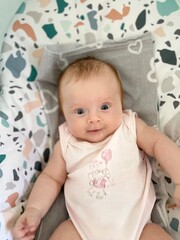 Smiling baby sitting in a colorful, patterned baby seat. The infant is dressed in a light...