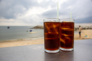 glass of iced cola sits on a table by the sea, with a cloudy sky backdrop. The drink's bubbles and...