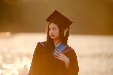 Asian woman graduate in cap and gown, posing by a golden, shimmering lake during sunset, signifying achievement and future aspirations.