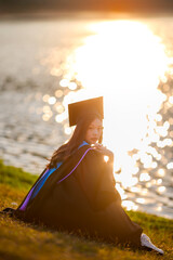 asian female graduate in a black academic gown and cap sits on the grass near a shimmering lake, looking back under warm golden sunlight during sunset.

