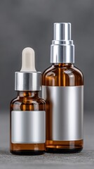 Naklejka premium Two amber glass cosmetic bottles with blank silver labels, one with a dropper and one with a spray, displayed on a gray background.