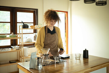 Smiling woman prepares coffee with pour over setup in a sunlit kitchen