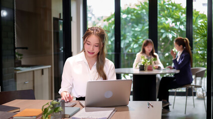 Asian Young Women Working on Laptop in Office with Colleagues Beautiful Adult Woman Concentrating on Computer Tasks in Modern Workplace with Natural Light