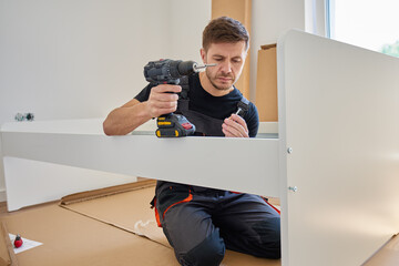 Focused man in bib overalls connects details of new bed in bedroom using electric drill. Assembling...