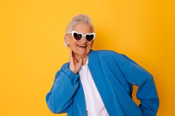 Senior woman in heart-shaped sunglasses and blue jacket smiling against a yellow background