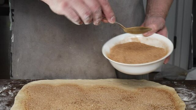 hands sprinkling cinnamon sugar mixture onto rolled out dough during preparation of cinnamon rolls on floured wooden surface, concept of bakery, dessert making, cooking class