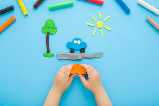 Little child boy hands making and creating car, tree and sun from colorful modeling clay on light blue table background. Pastel color. Closeup. Point of view shot. Top down view.