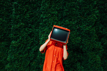 Woman posing outdoors with a retro CRT TV covering her head against a green hedge