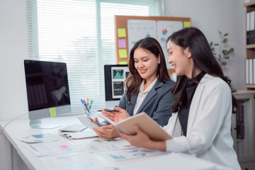 Two smiling Asian businesswomen are collaborating on a project, analyzing financial data with a calculator and documents, fostering teamwork and expertise in a modern office environment