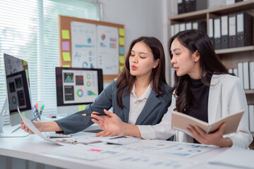 Two young businesswomen are examining financial charts and discussing data in a modern office, collaborating on a project and using a computer and documents