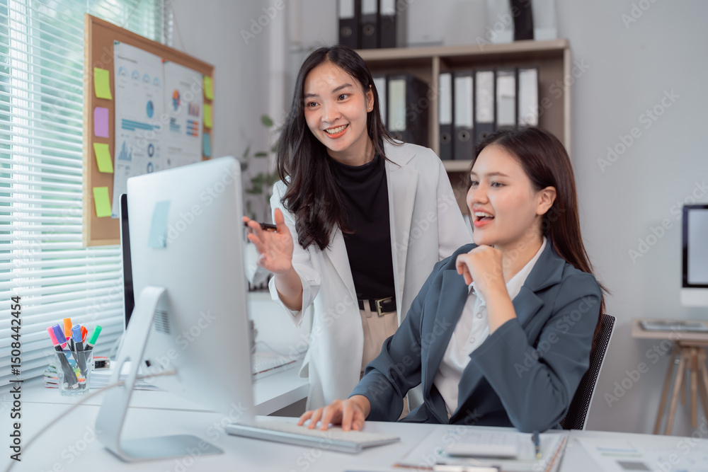 Poster two asian businesswomen are discussing a project while looking at desktop computer in a modern offic