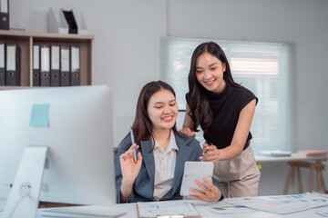 Two happy asian businesswomen discussing, brainstorming, collaborating on project using tablet and...