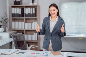 Asian businesswoman showing thumbs up with positive expression, standing near her desk with documents and charts, expressing success and agreement in modern office workspace