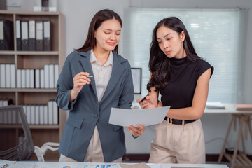 Two young businesswomen are discussing and analyzing financial data holding documents and pens,...