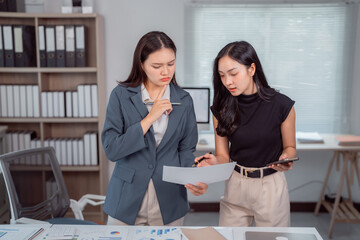 Two Asian businesswomen analyzing financial reports and discussing strategies to enhance...