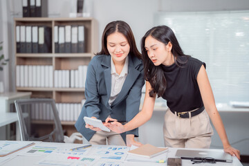 Two asian businesswomen are discussing and analyzing financial charts using calculator in a modern office, working together on a new project