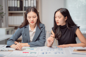 Two young businesswomen are discussing financial data and analyzing charts and graphs while working together at a table in a modern office, collaborating on a project and sharing their expertise