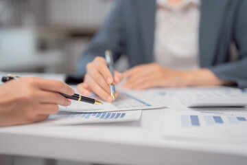 Two businesswomen are analyzing financial charts, pointing at data with pens, discussing statistics and using a calculator to make strategic decisions for their company