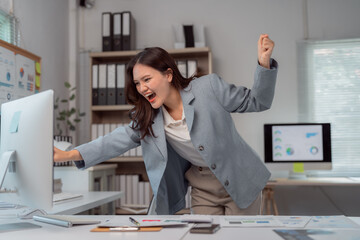Young Asian manager celebrating business success, raising arms in joy while looking at a desktop...