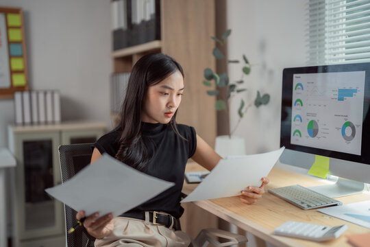 Focused Asian businesswoman comparing statistics document with data displayed on computer screen, working with financial graphs charts, analyzing marketing research results in office