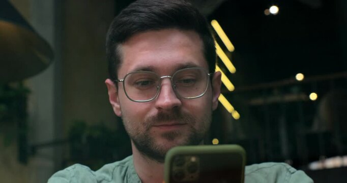 View through the window of handsom man using smartphone at coffee shop, reading social media internet, typing text or shopping online.