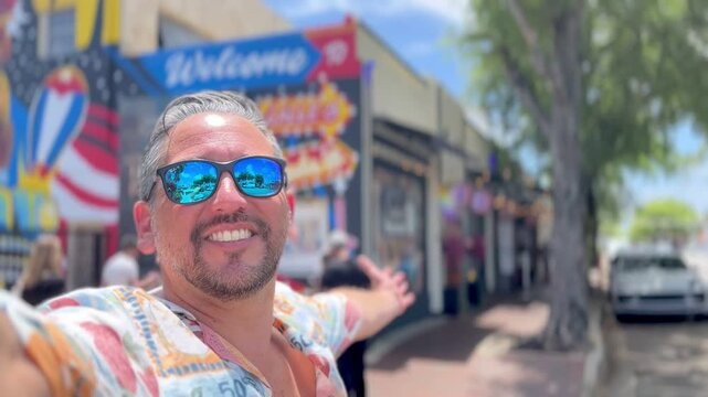 Mid adult latin travel tourist male wearing sunglasses, smiling joyfully while taking a video selfie with outstretched arms in Calle Ocho, Little Havana, enjoying a vibrant urban atmosphere
