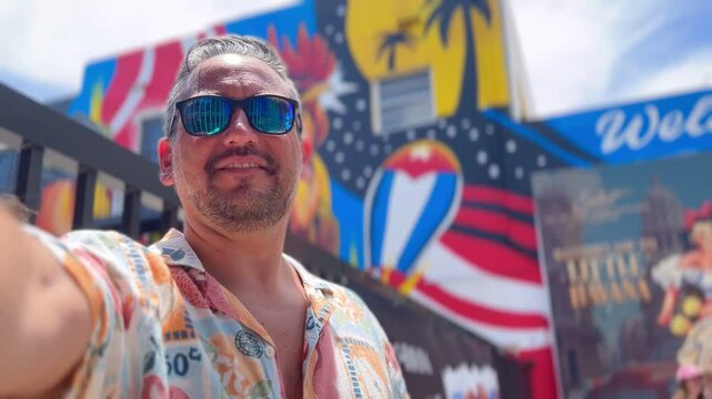 Mid adult latin travel tourist male wearing sunglasses, smiling joyfully while taking a video selfie with outstretched arms in Calle Ocho, Little Havana, enjoying a vibrant urban atmosphere