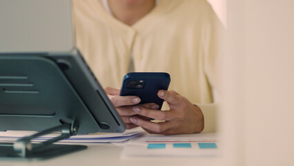 Close-up of female hands using smartphone while working at home. Digital communication, multitasking, and remote work with tablet, document, and modern workspace tools.