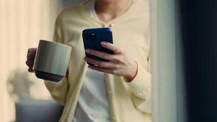 Close-up of woman holding smartphone and coffee mug at home.