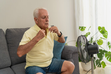 Sweating retired man sitting on sofa cooling with electric fan during summer heat at home