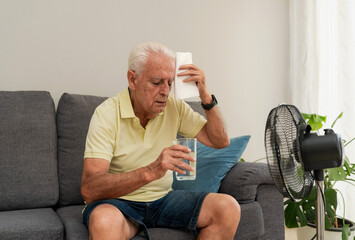 Senior man sweating and holding glass water while sitting on sofa in front of electric fan at home