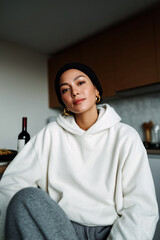 Smiling woman in hoodie relaxing at sunset indoors, cozy modern lifestyle portrait