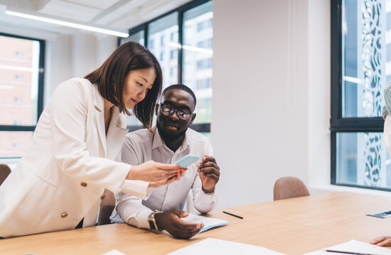 Asian businesswoman showing smartphone content to African American male colleague, both focused on screen while reviewing project details at wooden desk in open-plan office