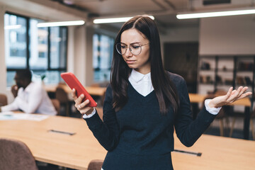 Young brunette woman in glasses looking confused while reading phone message, standing in office...