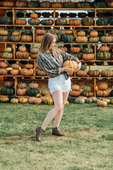 young woman stands at pumpkin festival, holding large orange and green pumpkin. She is surrounded by rows of pumpkins and a wooden display. The autumn farm setting captures the harvest season vibe.