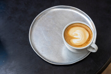 A latte with a heart design in the foam sits on a silver tray on a dark table...