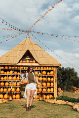 young woman stands at pumpkin festival, holding large orange and green pumpkin. She is surrounded by rows of pumpkins and a wooden display. The autumn farm setting captures the harvest season vibe.