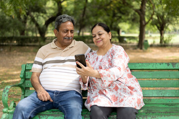 Fototapeta premium Indian elder or senior happy couple using mobile phone at the park while sitting on bench