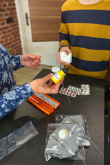 Unrecognizable woman holding iodine antiseptic and medical supplies showing to teenager boy at home, with face masks, pills and band aids visible on kitchen counter, teaching safety and health.
