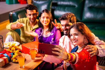 Smiling and clicking festive selfie while holding Diwali sweets with family and friends at home